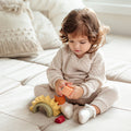 A toddler with curly hair plays with the Nuby US One & Done Bunny Basket Bundle, a sensory play set featuring colorful arches and cylinders, while sitting on a beige sofa with cozy cushions and an Easter basket in the background.
