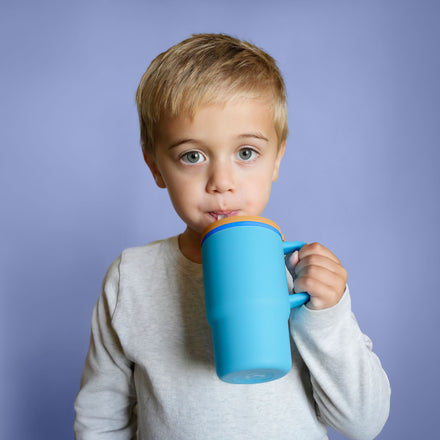 A young boy with blond hair and blue eyes drinks from the Silicone Tiny Tumbler in Electric Blue, featuring a large spill-proof design with a silicone straw. He is wearing a light gray shirt and standing in front of a solid lavender background.