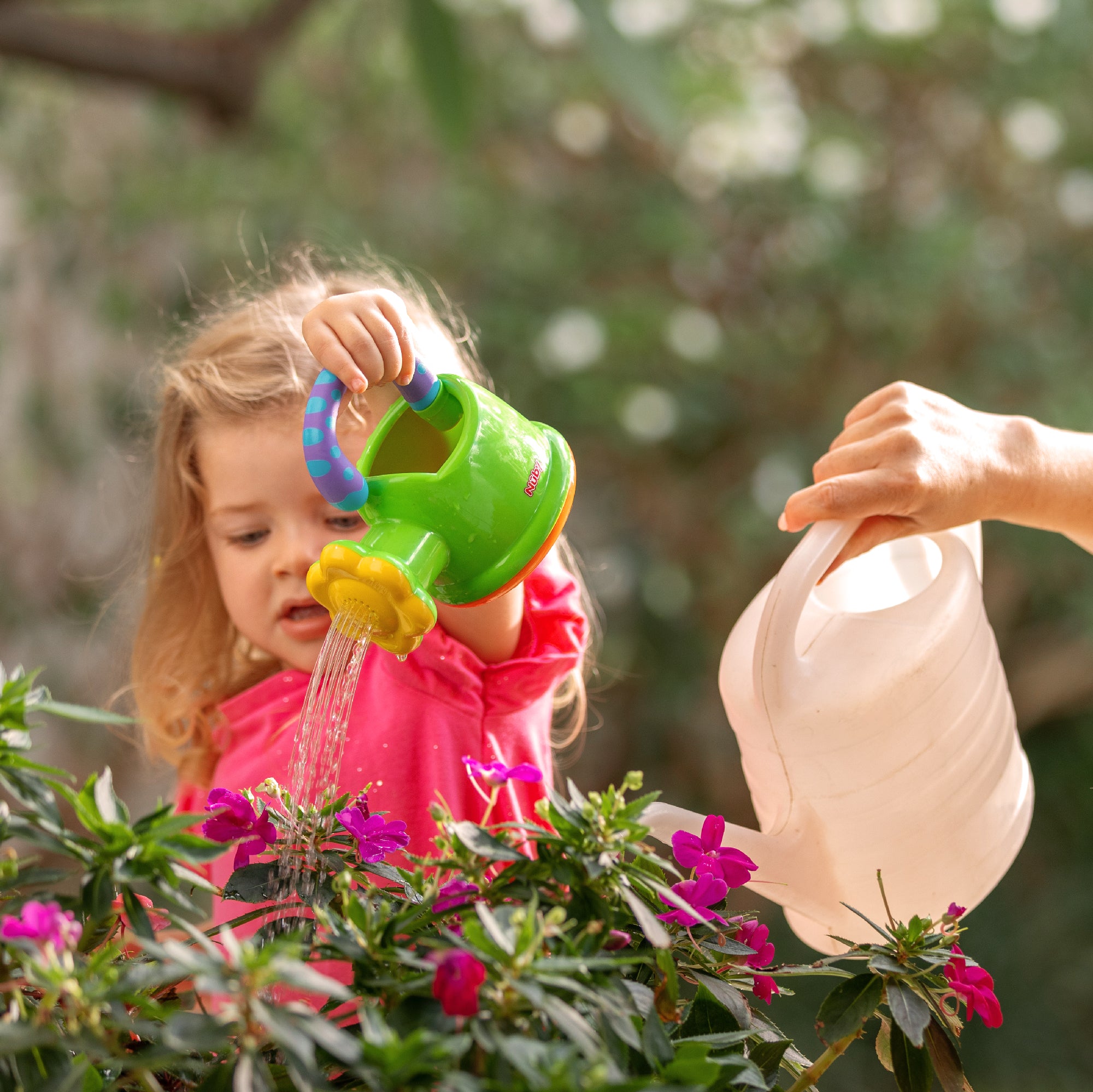A young child in a pink shirt uses the Nuby US One & Done Bunny Basket Bundle's green toy watering can to water blooming pink flowers, while an adult pours water from a white can. Lush green foliage fills the background.