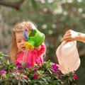 A young child in a pink shirt uses the Nuby US One & Done Bunny Basket Bundle's green toy watering can to water blooming pink flowers, while an adult pours water from a white can. Lush green foliage fills the background.