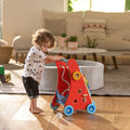 A toddler with curly hair plays with a colorful activity walker in a sunlit living room, standing on a wooden floor near a white rug and modern Montessori-inspired furniture.