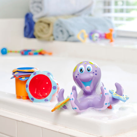 The Nuby Splish Splash Stacking Cups – 5 pack, in bright colors, sit on the bathtub edge with a smiling purple octopus bath toy. Foam rings and towels are visible, making bath time fun and perfect for baby development.