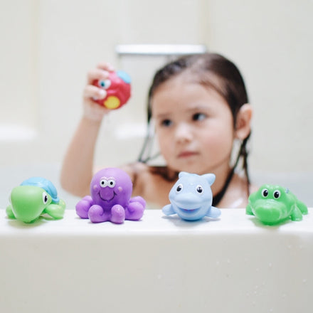 A young child in a bathtub holds up a red Nuby Little Squirts Bath Squirters toy, while four colorful animal squirters from the 10-pack—including a turtle, octopus, dolphin, and alligator—are lined up on the tub's edge.