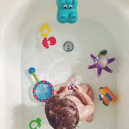 A small child enjoys bath time with the Nuby Splash n' Catch Bathtime Fishing Set, surrounded by colorful toys like a blue hippo and purple octopus—perfect for encouraging hand-eye coordination during play.
