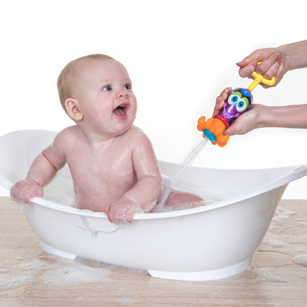 A smiling baby sits in a white bathtub with suds, looking up as an adult sprays water with the colorful Nuby Squid Squirter Water Squirter, a fun bathtime toy shaped like a playful cartoon squid.