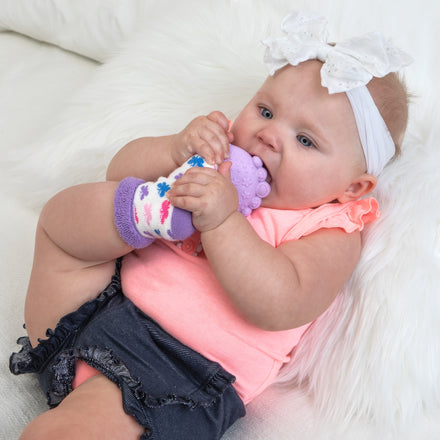 A teething baby in a white headband, pink shirt, and dark shorts lies on a white furry surface, chewing on the Nuby Soft Teether Sock Set with Travel Bag, which features a soothing silicone teether attached for relief.