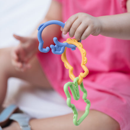 A young child in a pink dress holds Nuby’s Animal Click Links, a baby toy designed to support hand-eye coordination, while sitting on a soft surface.