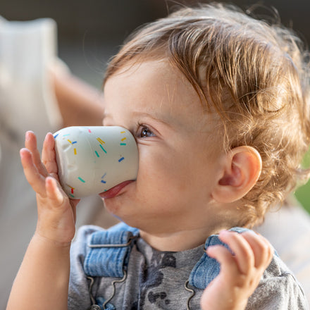 A young child in a gray shirt and denim overalls practices open cup drinking, sipping from the Nuby US Baby's First Mini Sipper Training Cup decorated with colorful sprinkles.